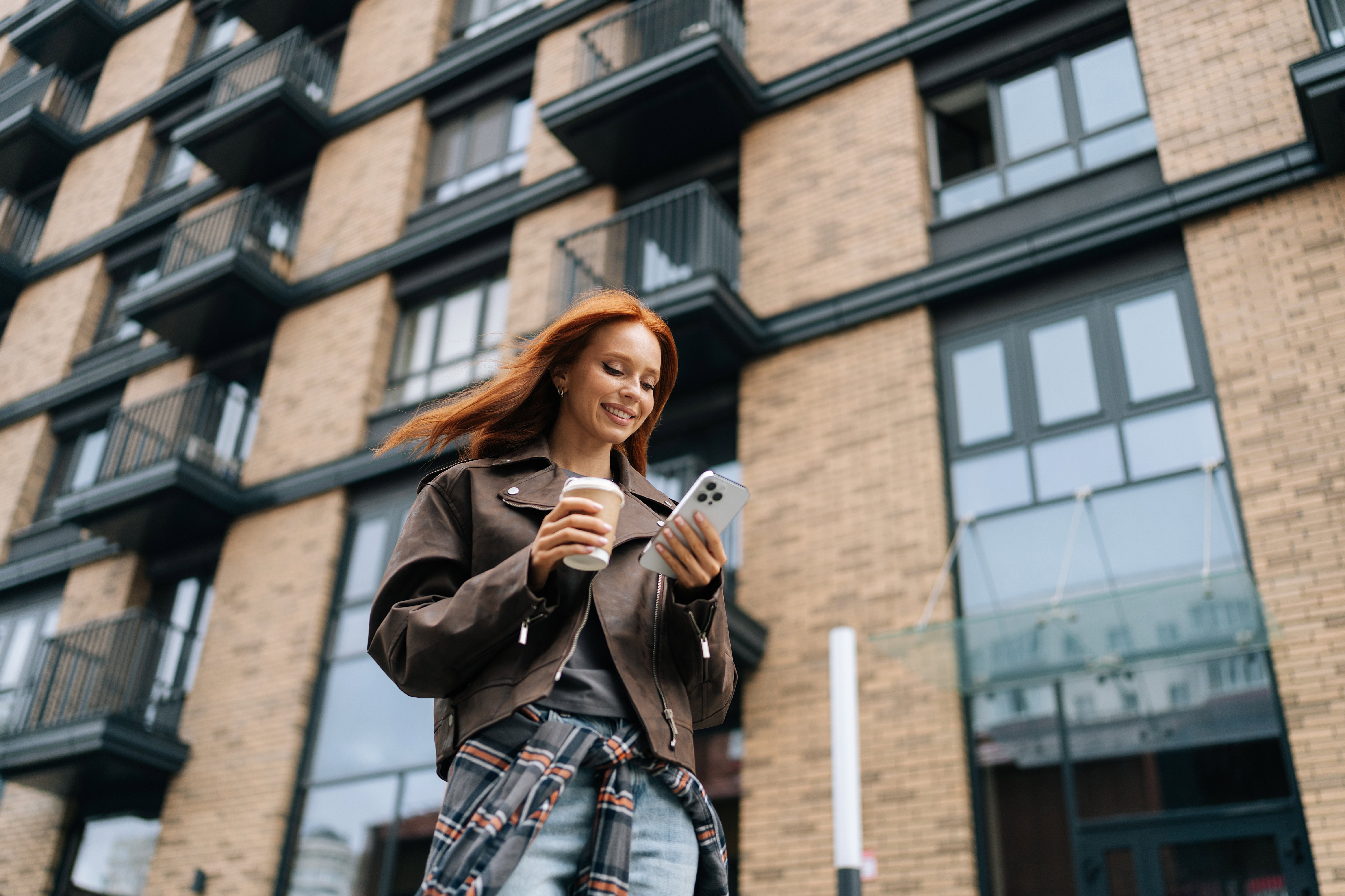 Low-angle view of stylish cheerful young woman standing on city street while drinking coffee and browsing smartphone with smile on face