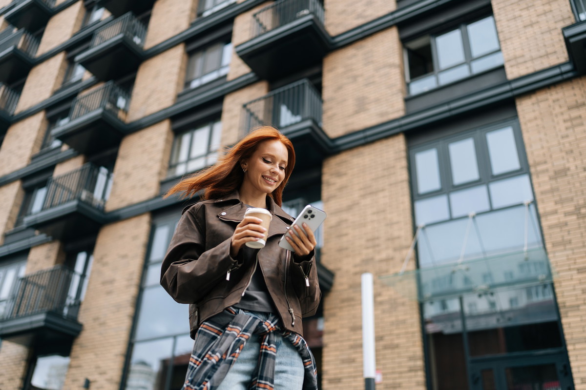 Low-angle view of stylish cheerful young woman standing on city street while drinking coffee and browsing smartphone with smile on face