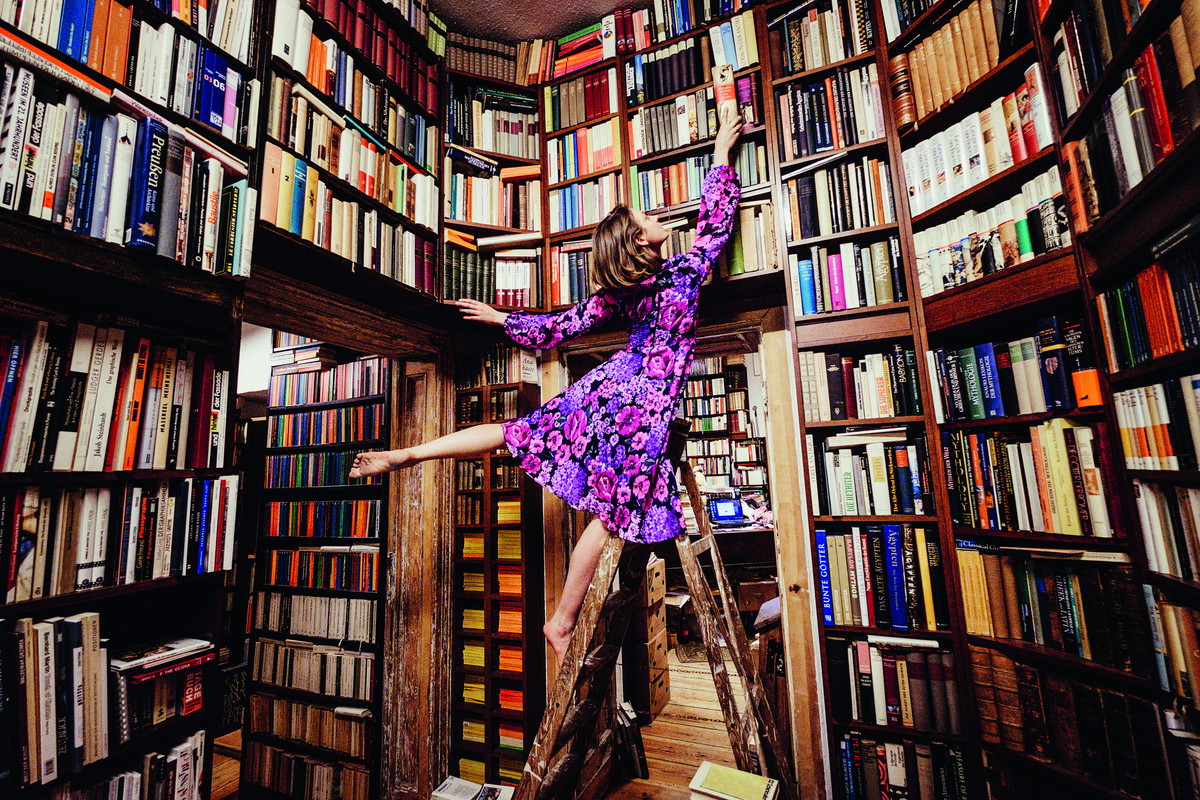 Carefree woman on ladder reaching for book in library