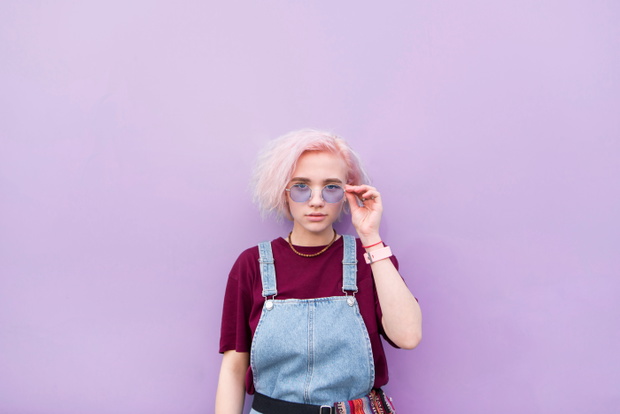 Stylish girl with pink hair and sunglasses is a blue background and looks at the camera. Portrait of a stylish girl against a background of blue wall