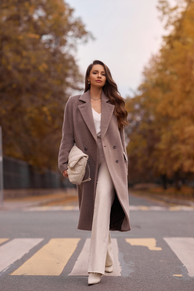 Elegant brunette woman with long wavy hair wearing grey wool coat, holding white handbag and walking at city street on autumn day. Portrait against yellow leaves
