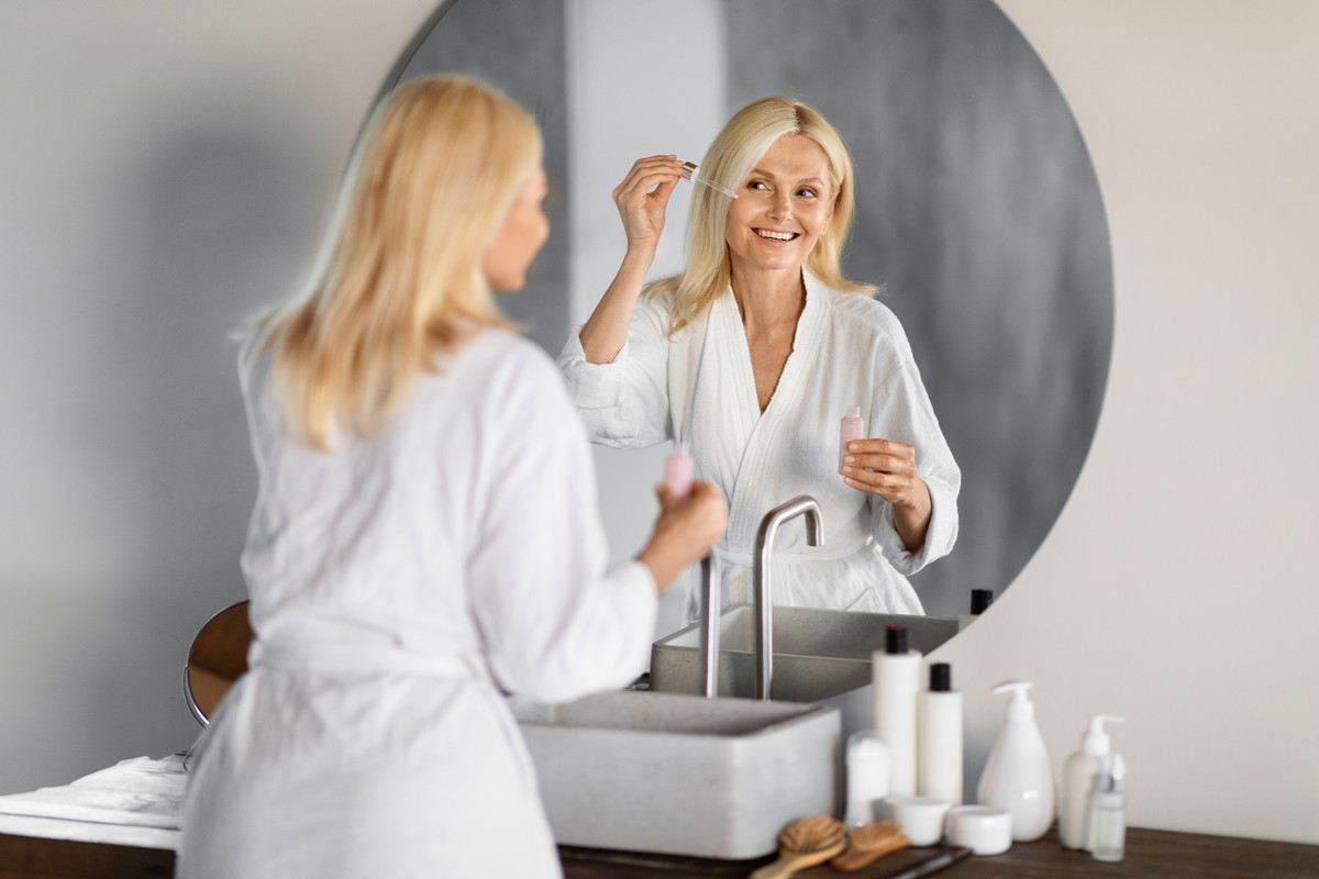 Elegant,Senior,Woman,In,White,Bathrobe,Applying,Facial,Serum,,Looking