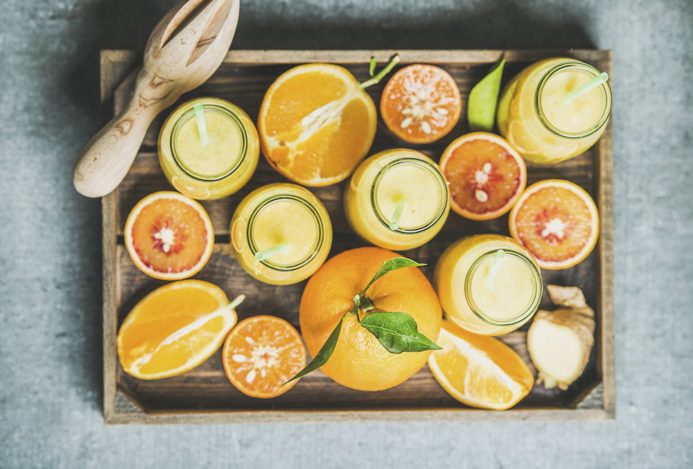 Healthy yellow smoothie with citrus fruit and ginger in bottles in wooden tray over grey concrete background