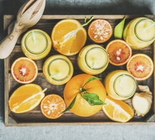 Healthy yellow smoothie with citrus fruit and ginger in bottles in wooden tray over grey concrete background