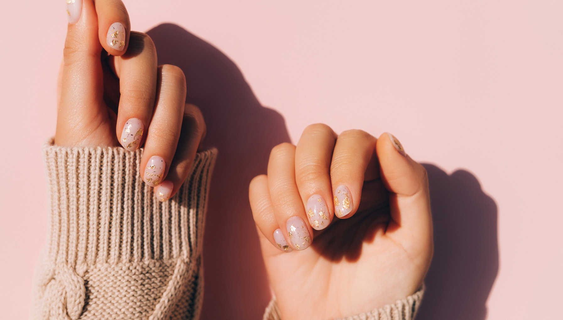 Glowing hands in sweater with knitted sleeves and with nude color manicure with gold particles on pink table. Trendy colors of the year