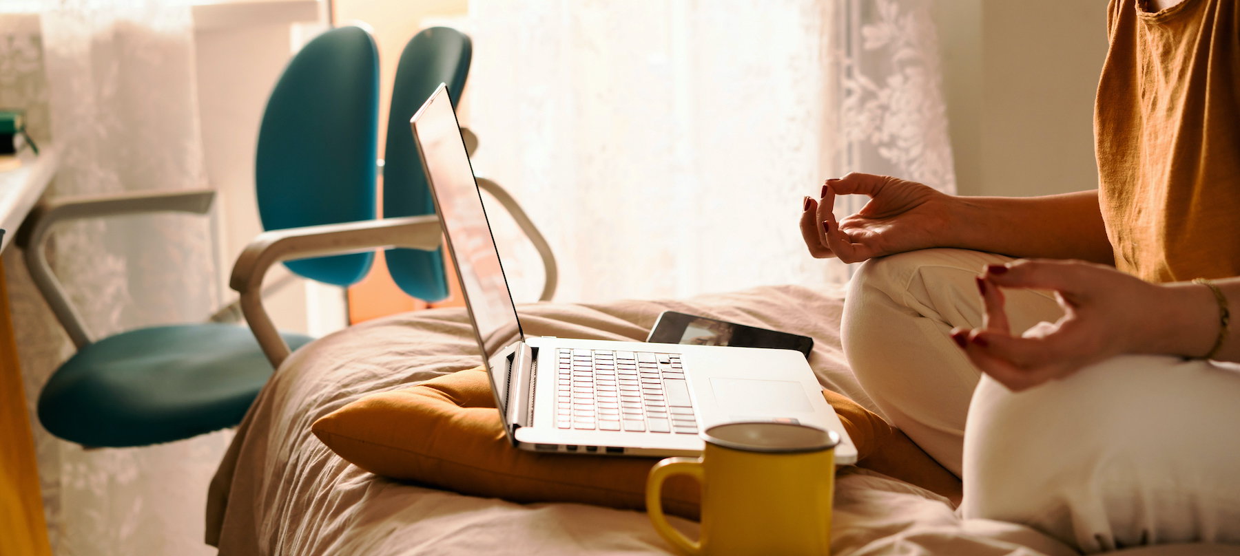 A middle-aged woman in white jeans and a yellow sweater sitting on the bed in a yoga pose in front of a laptop and a cup of coffee