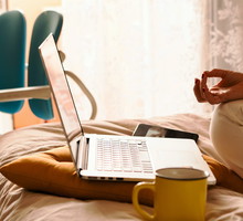 A middle-aged woman in white jeans and a yellow sweater sitting on the bed in a yoga pose in front of a laptop and a cup of coffee