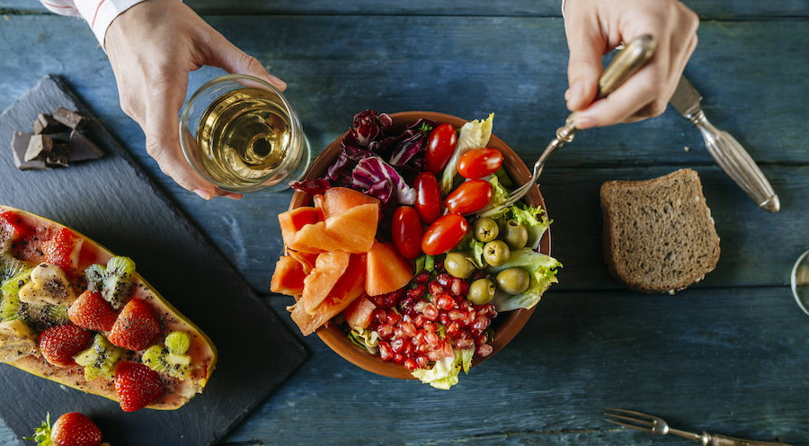 Close-up of woman's hands eating salad wit tomato, pomegranate, papaya and olives, papaya with fruits and with wine glass
