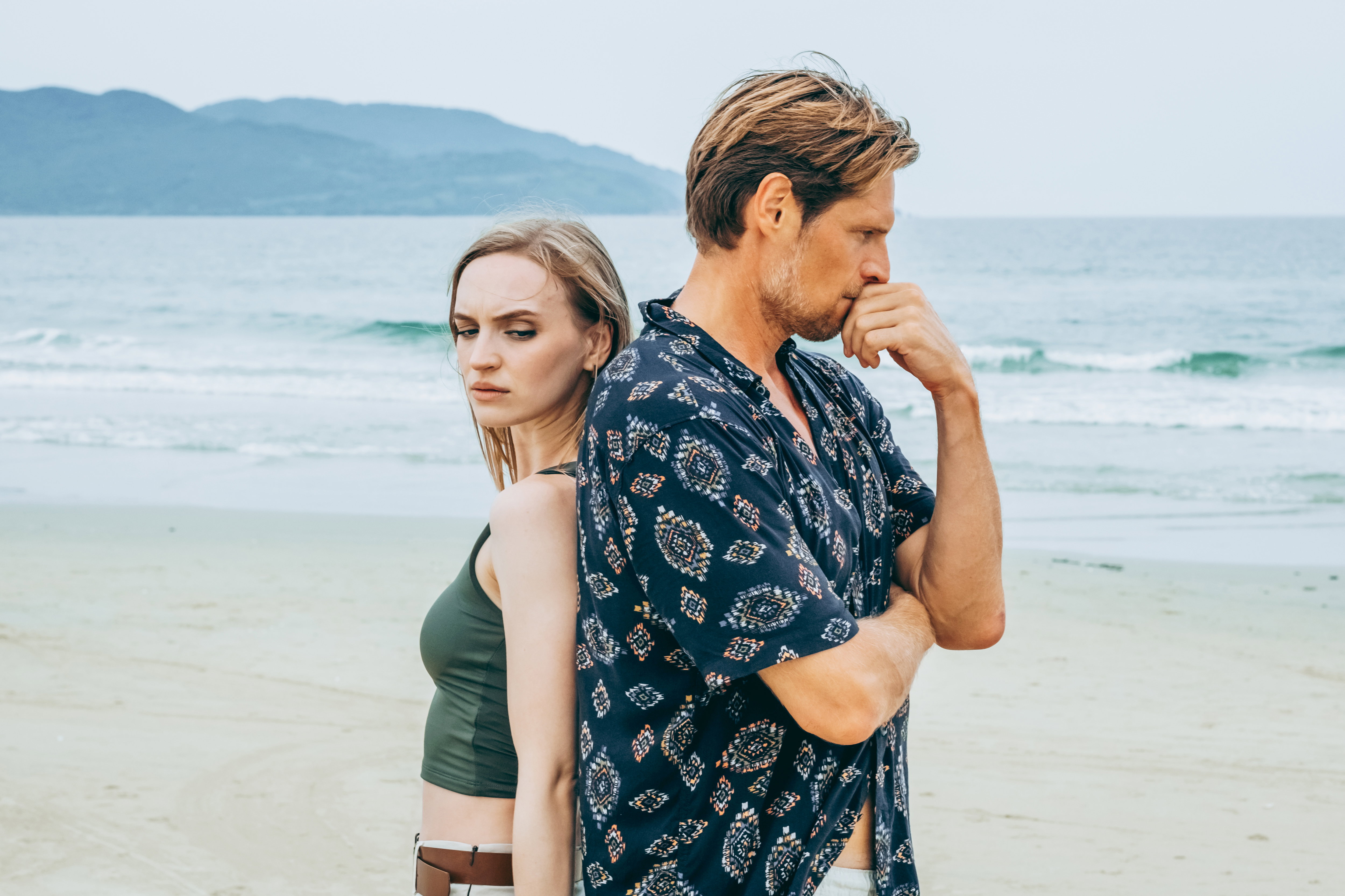 Pensive couple standing back-to-back on tranquil beach by the ocean