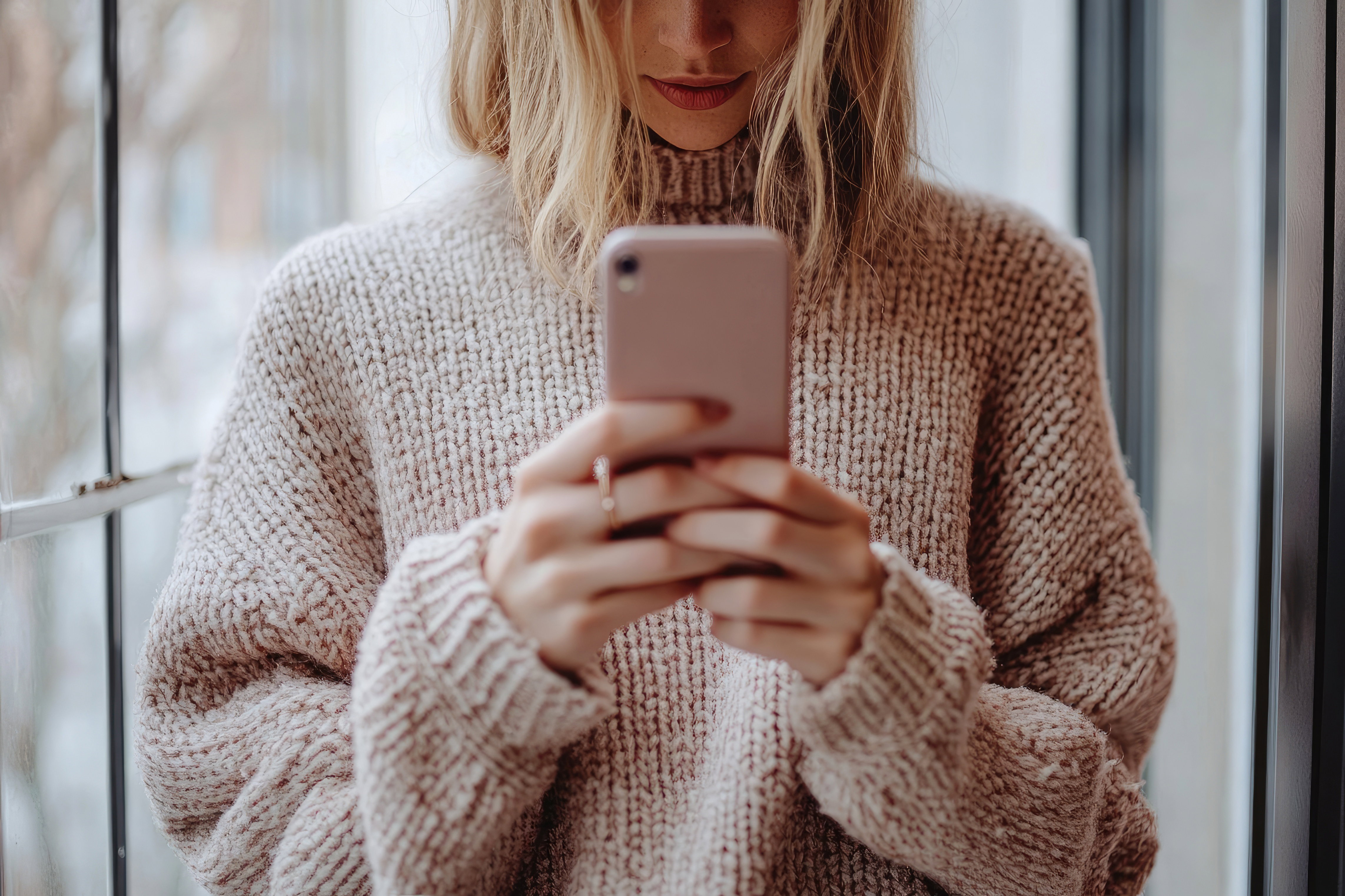 Woman wearing cozy sweater using smartphone near window