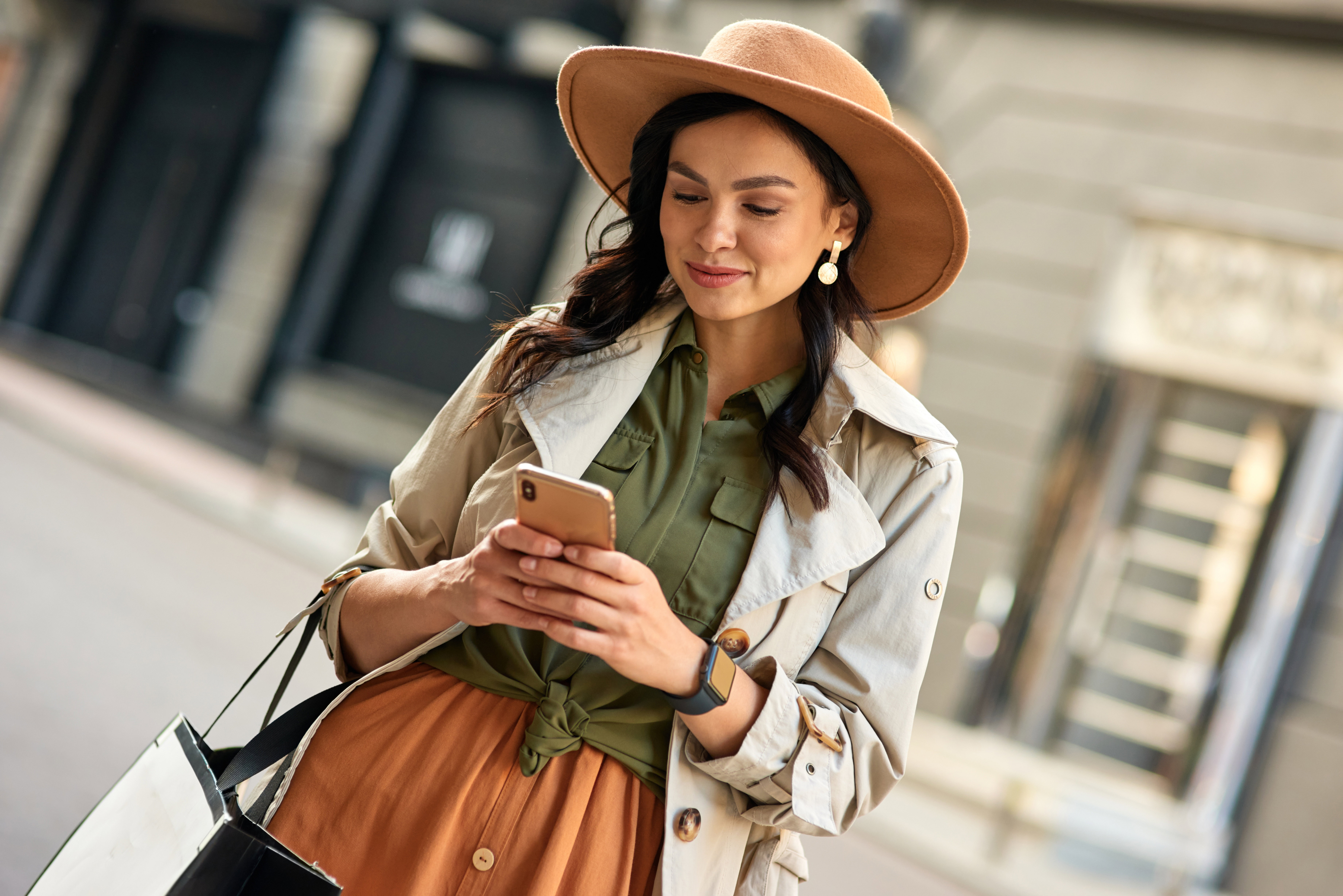 Chatting with friend. Young beautiful stylish woman wearing autumn coat and hat using her smartphone and smiling while standing on city street