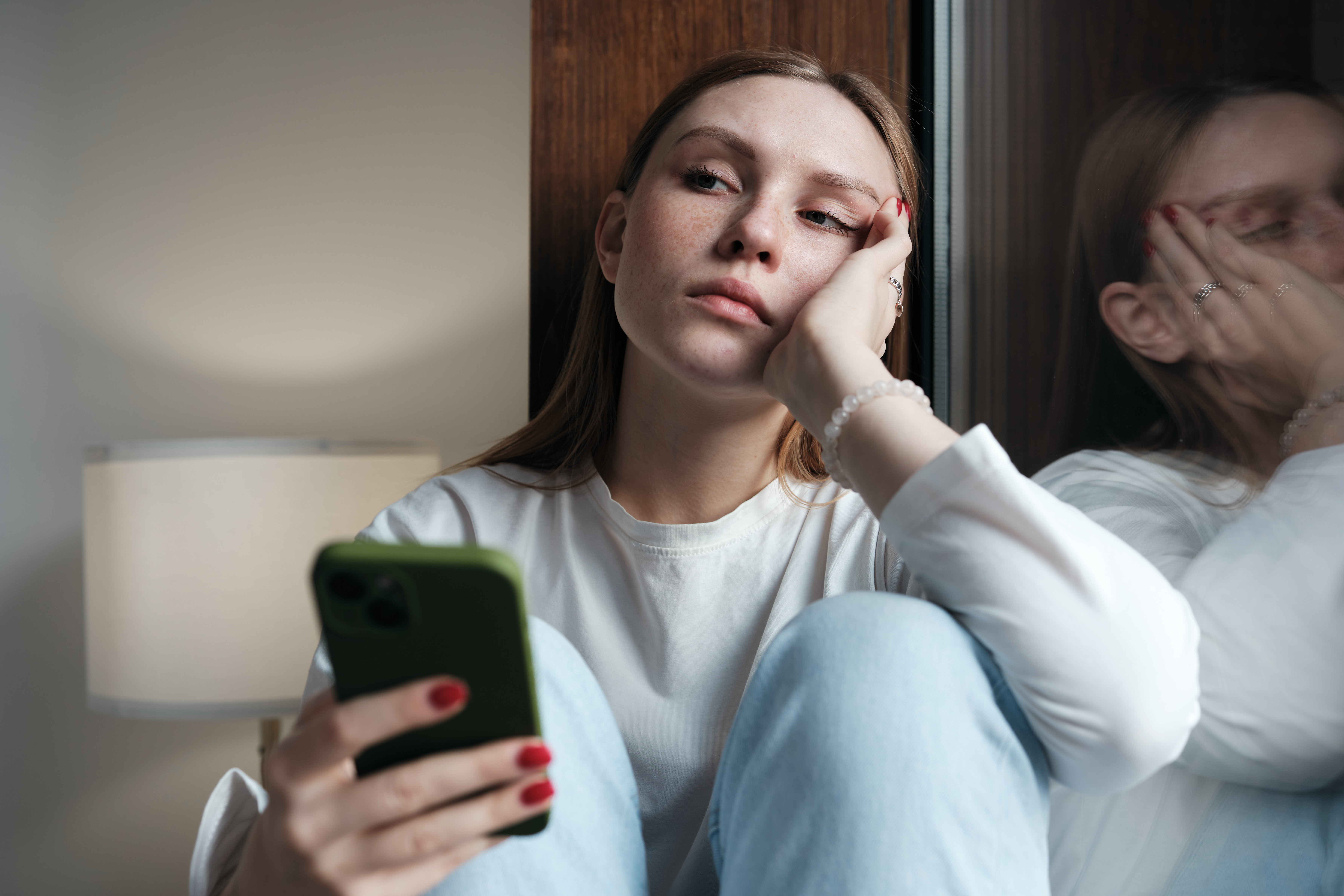 Upset redhead teen girl sitting by window looking at phone waiting call or message
