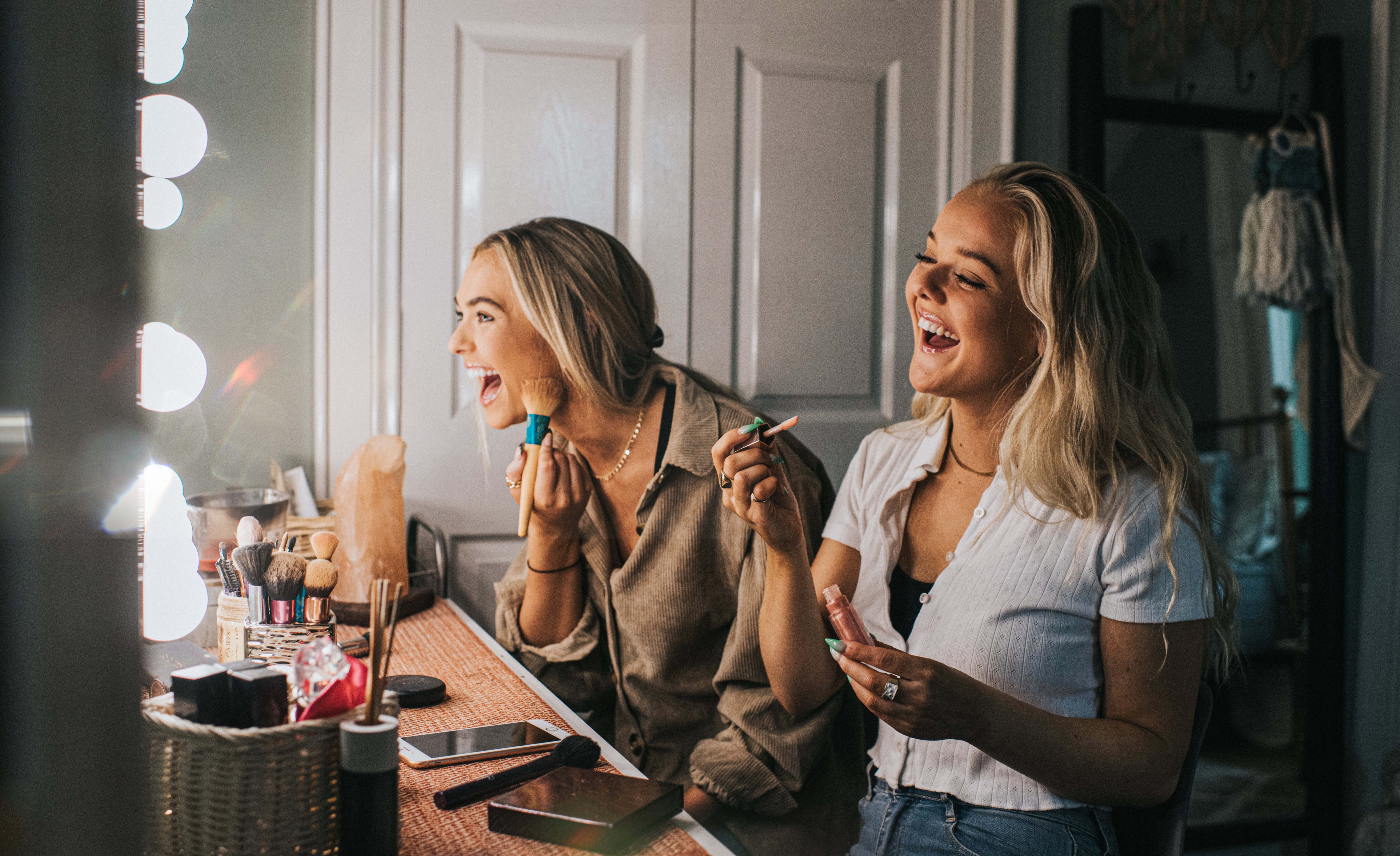 Two young woman sit in front of an illuminated mirror and apply make-up