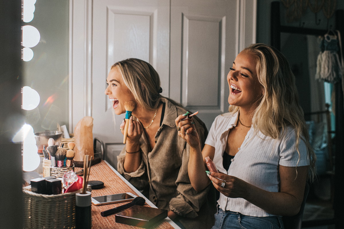 Two young woman sit in front of an illuminated mirror and apply make-up