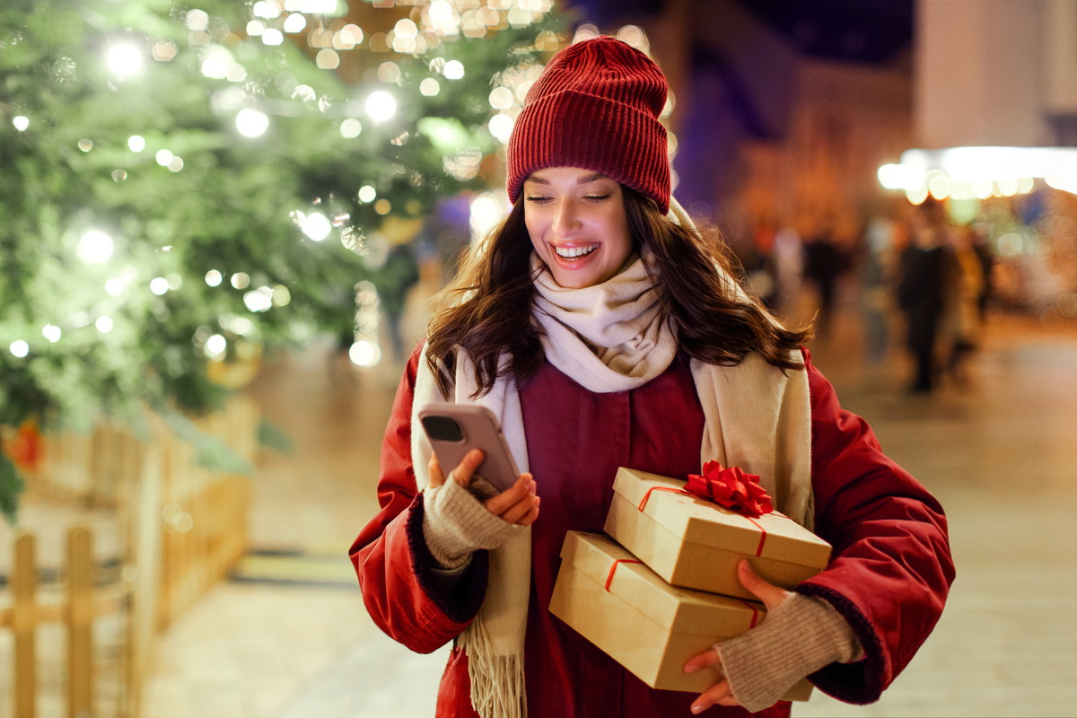 Modern holiday shopping. Happy lady holding gift boxes and using smartphone while standing outdoors near Xmas tree and festive decorations