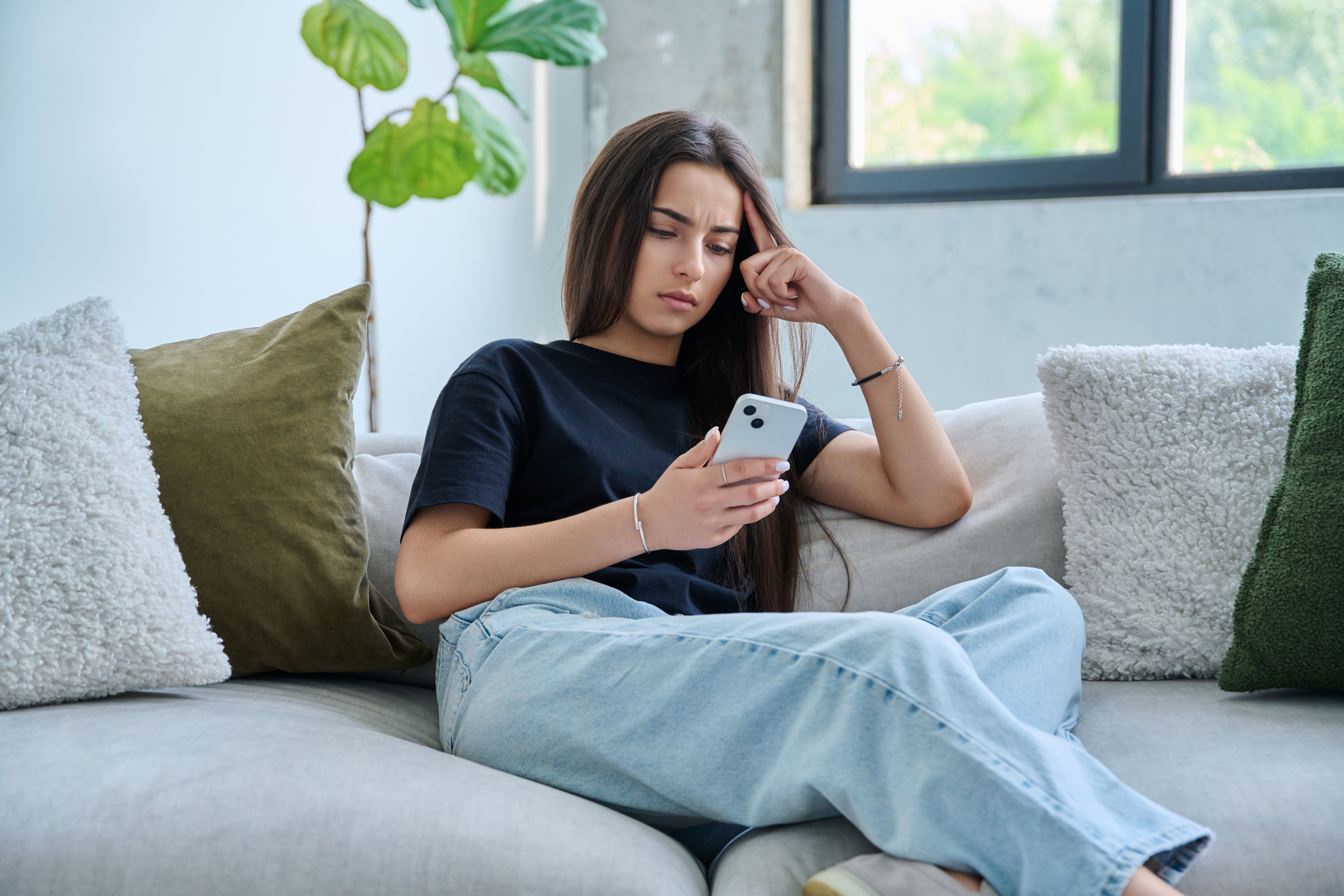 Upset sad young woman looking at smartphone sitting on couch at home