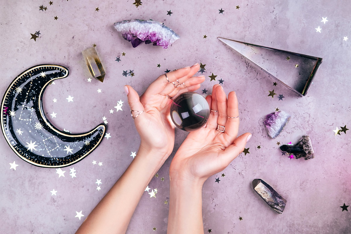 Magic crystals for rituals. Hands with rings on fingers are holding crystal ball near esoteric set on concrete gray background with many stars sequins. Flat lay style