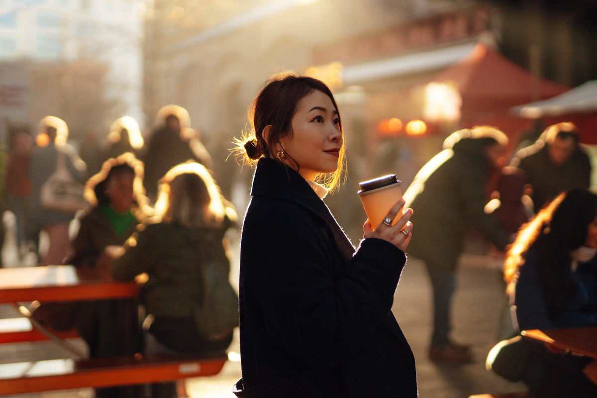 Young Asian woman drinking coffee at local street food market with moody sunlight