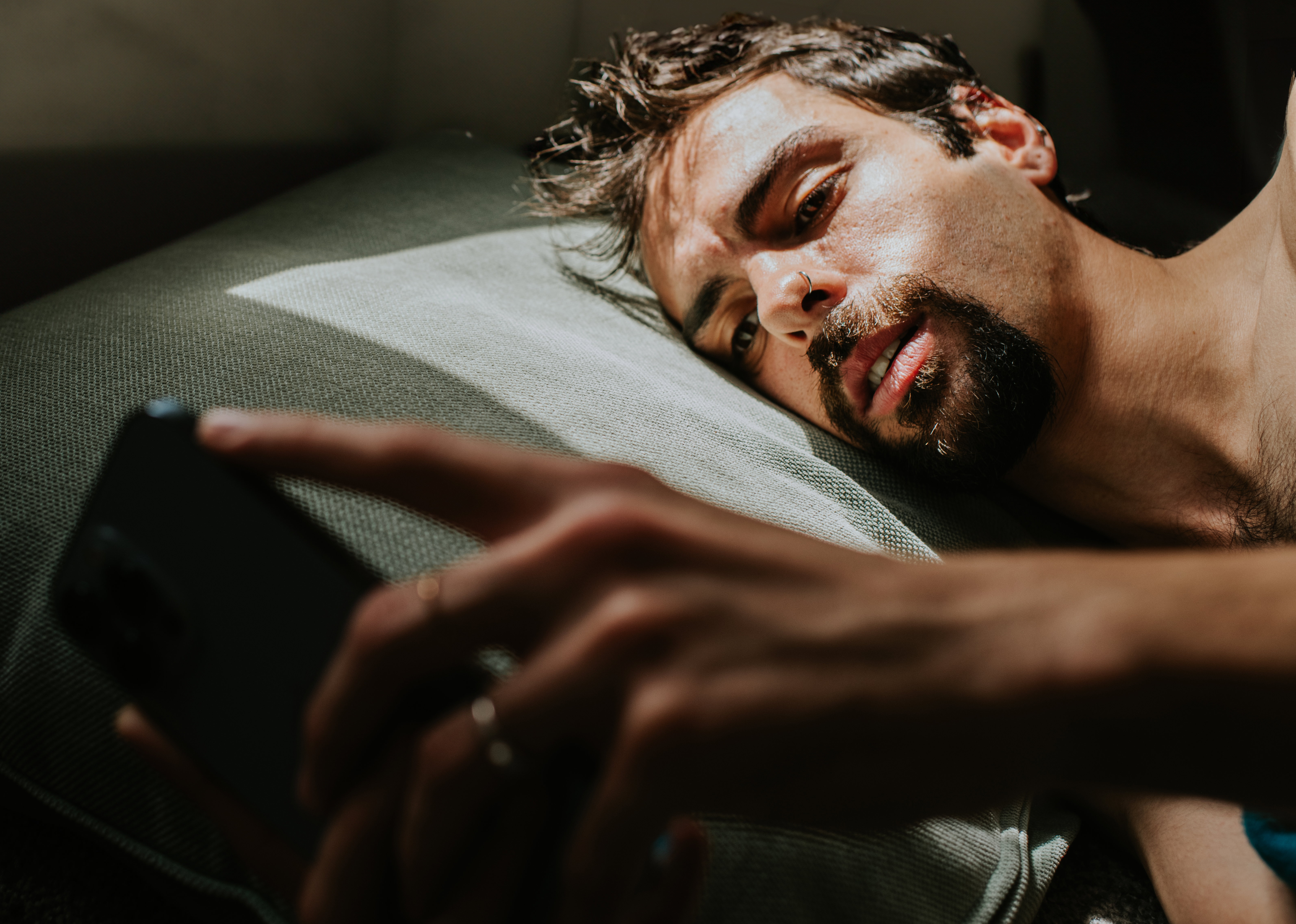 A man lies on his side in bed, with a blank expression, looking at a smart phone
