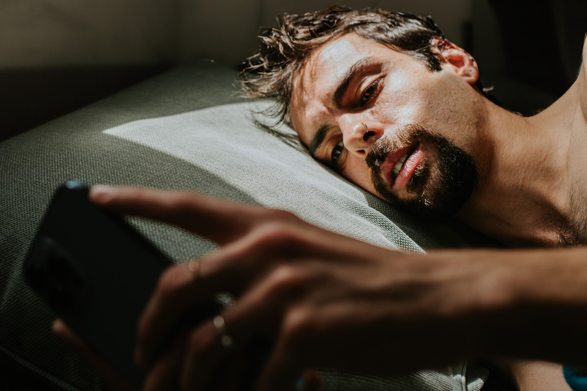 A man lies on his side in bed, with a blank expression, looking at a smart phone