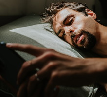 A man lies on his side in bed, with a blank expression, looking at a smart phone