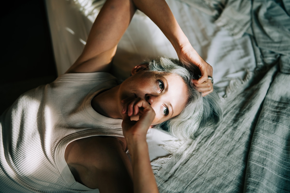 A middle-aged woman with gray hair lies on her back in bed, illuminated by bright sunlight and shadow.