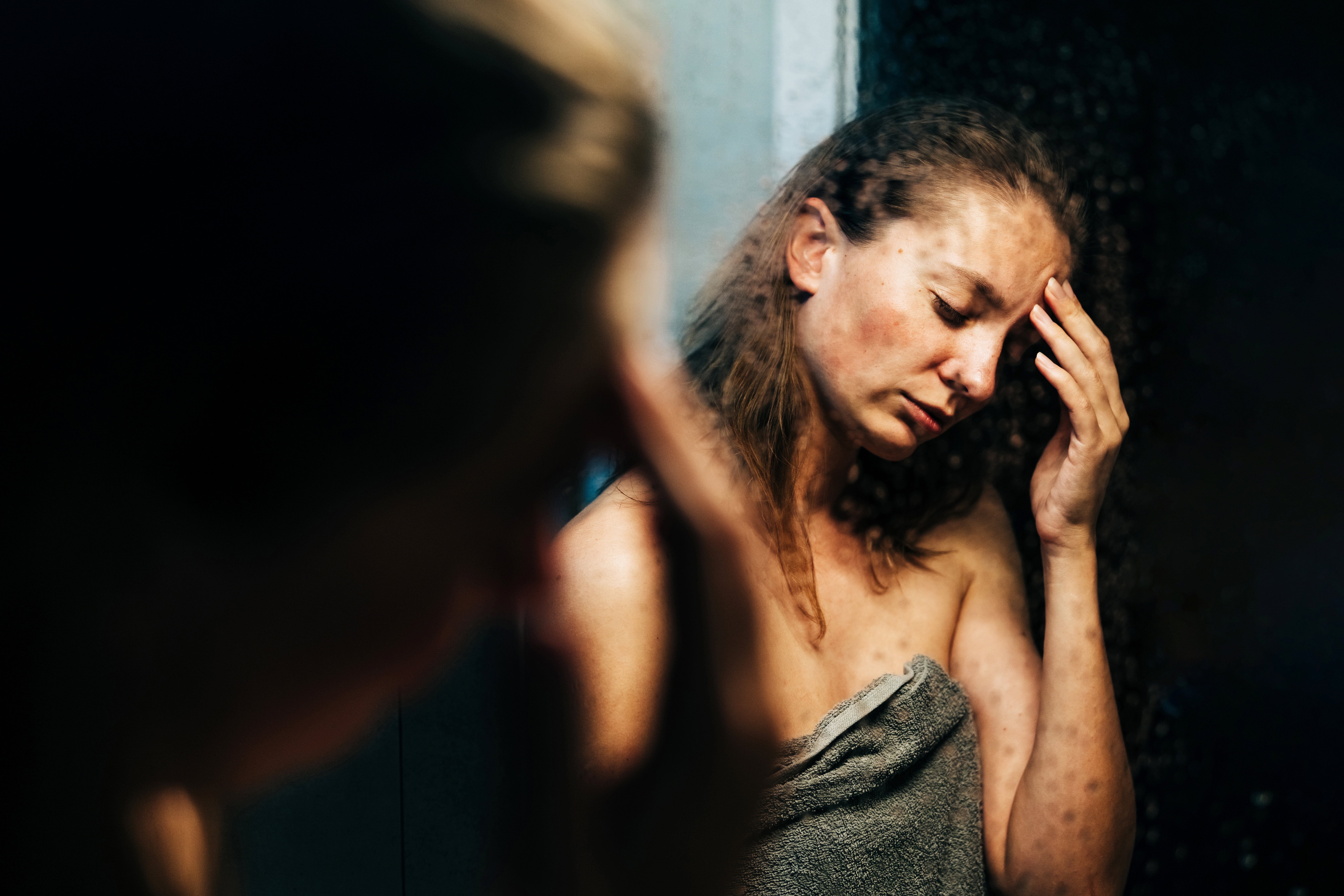 A woman wrapped in a towel touches her forehead while looking in a mirror with a tired expression.