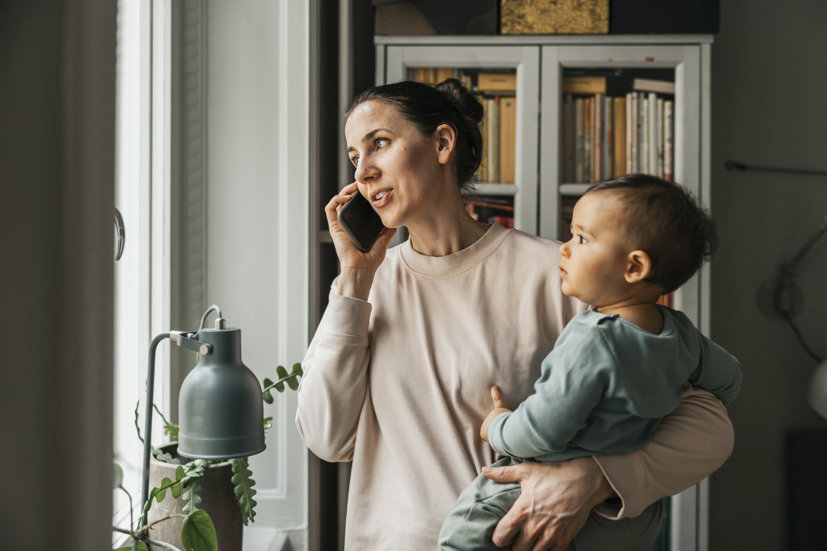 Mother carrying baby boy in arm while talking on smart phone at home