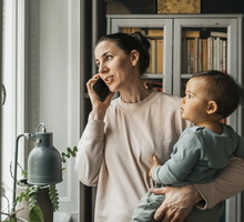 Mother carrying baby boy in arm while talking on smart phone at home