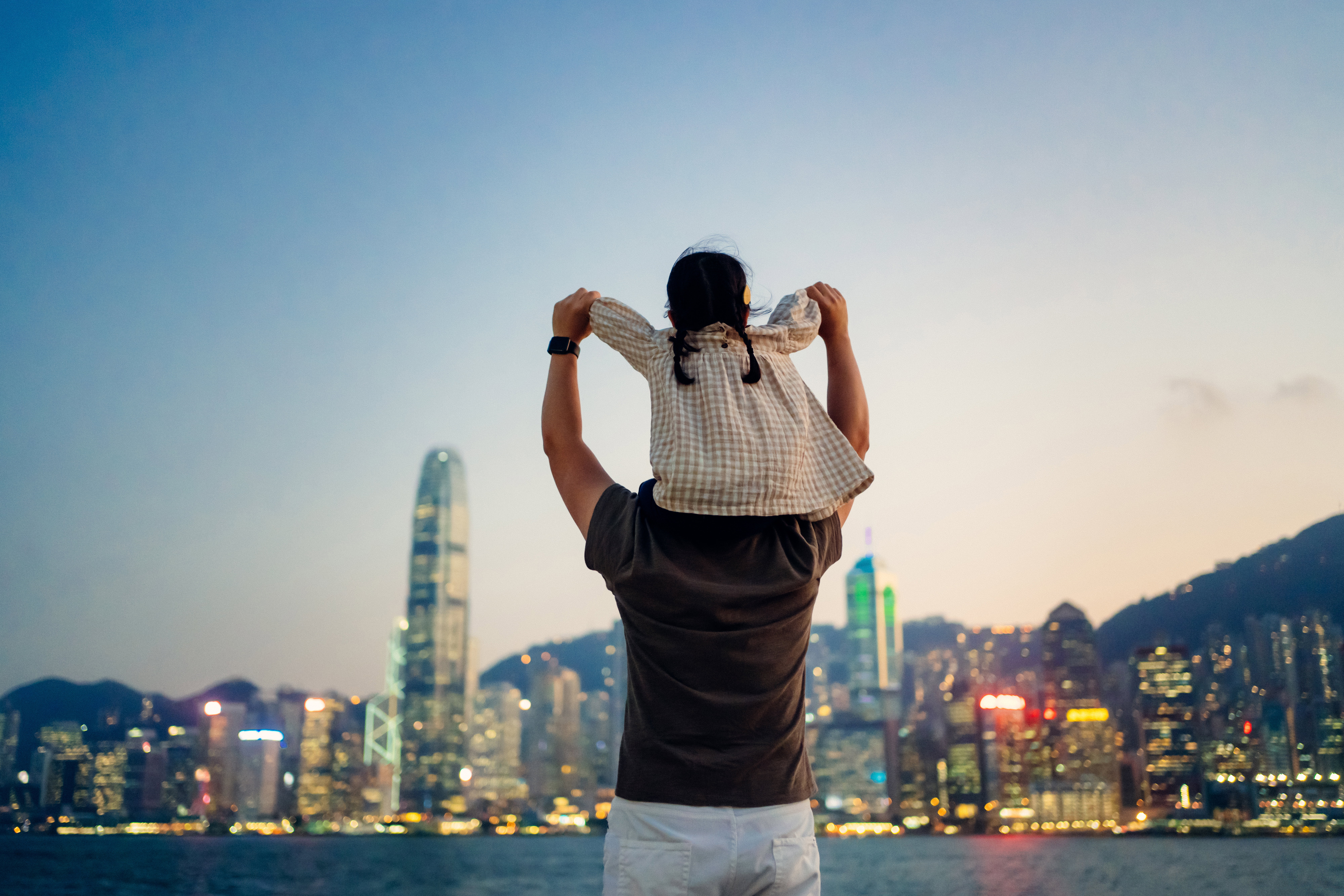 Loving young Asian father carrying little daughter on shoulders, looking over illuminated urban city skyline against the promenade of Victoria harbour at sunset. Enjoying father and daughter bonding time together