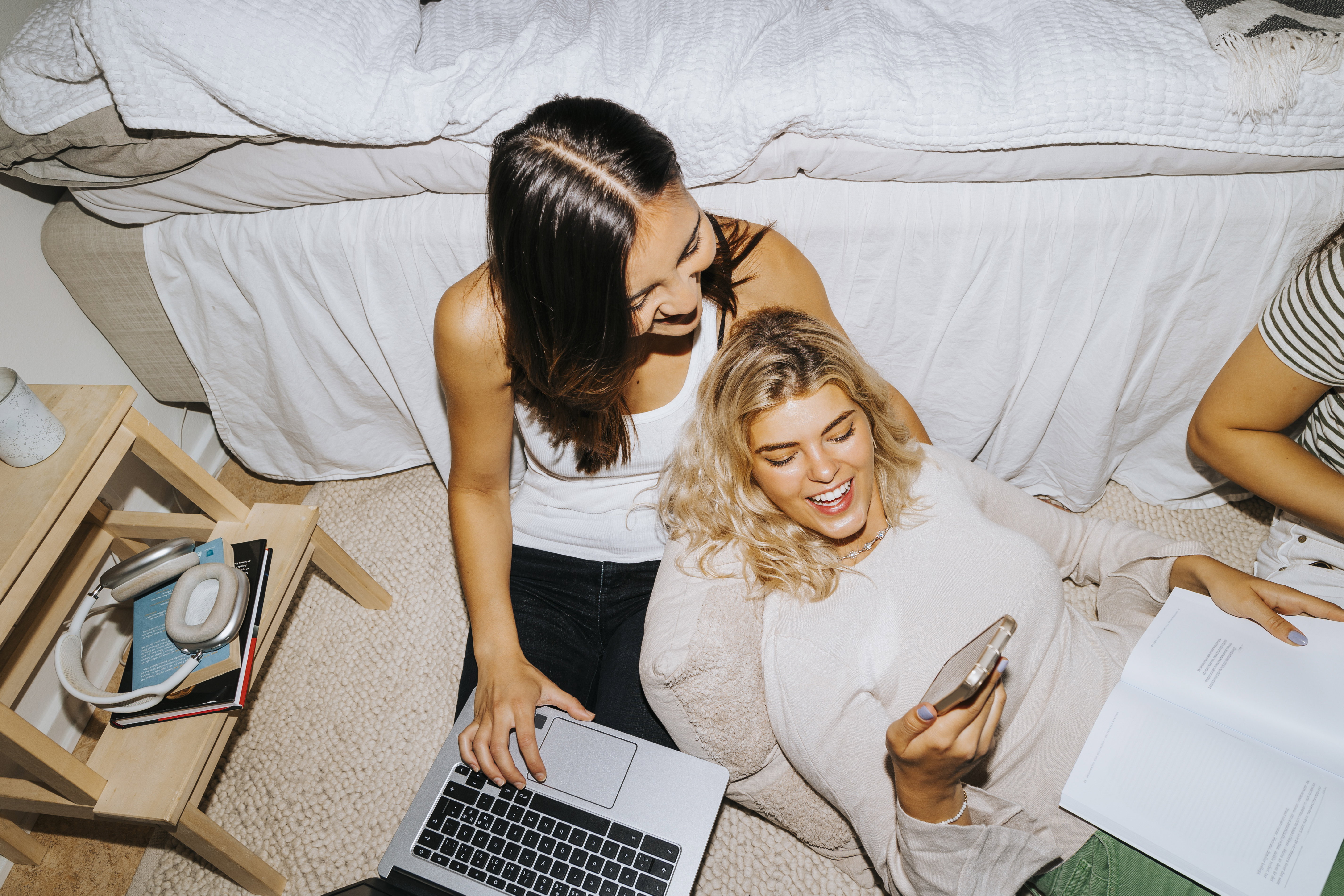 High angle view of smiling woman sharing smart phone with female friend sitting by bed in college dorm