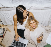 High angle view of smiling woman sharing smart phone with female friend sitting by bed in college dorm