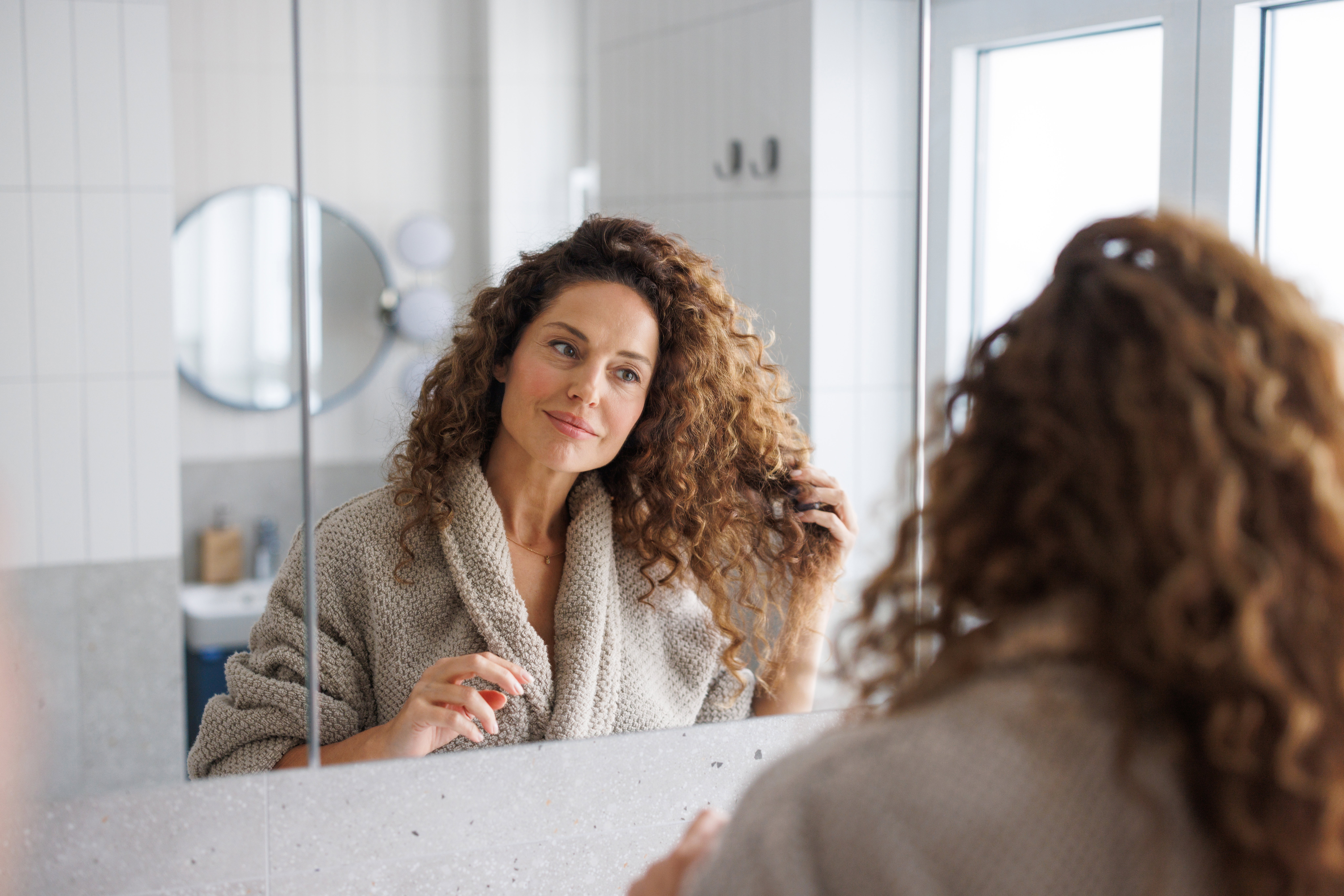 Thoughtful mid-adult woman with curly hair looking at herself in the bathroom mirror
