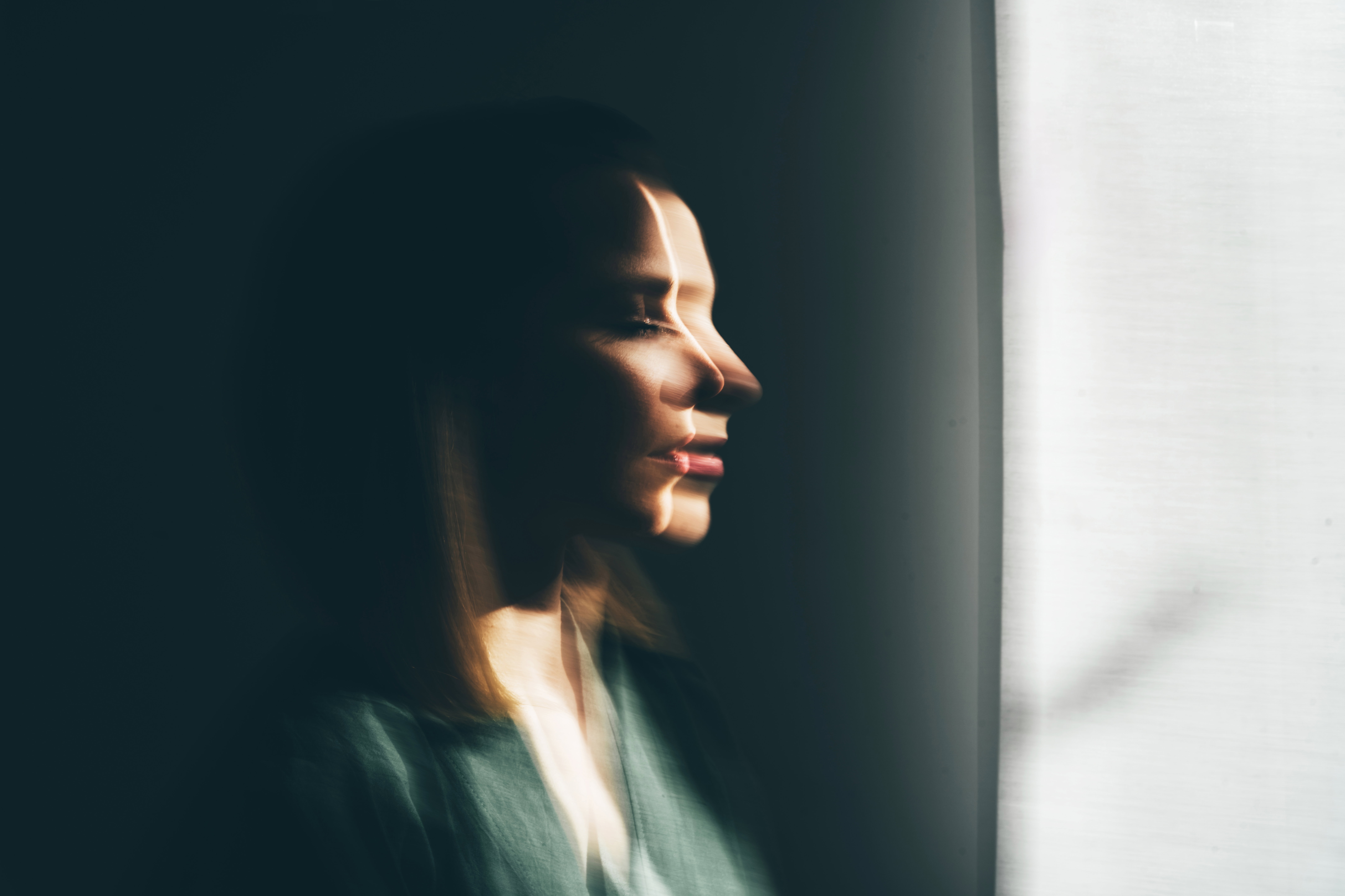 A conceptual portrait of a woman with a double exposure effect, standing in shadows with a slit of light on her face.