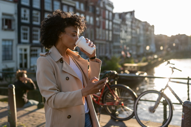 Woman drinking coffee using smartphone on amsterdam canal bridge at sunset, lifestyle travel connection concept