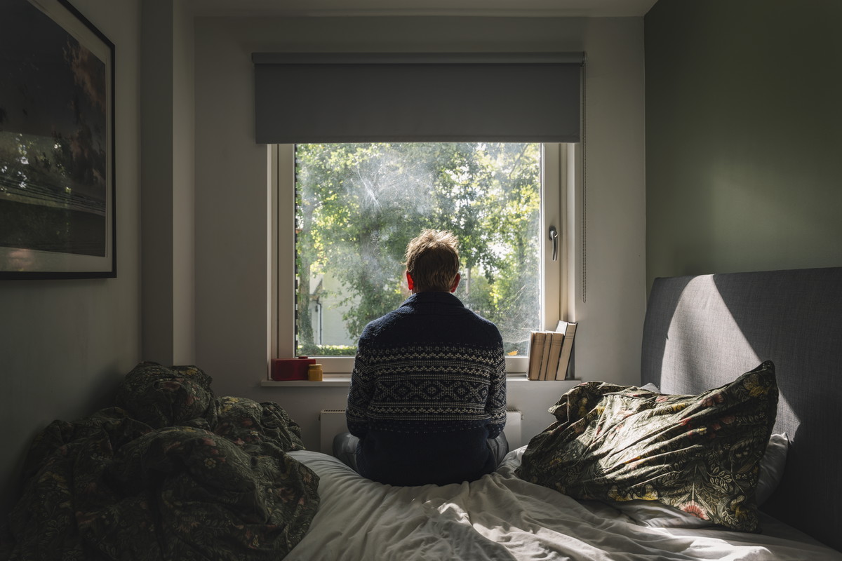 Rear view of a depressed man looking out of his bedroom window on a sunny day - negative emotion