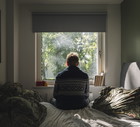 Rear view of a depressed man looking out of his bedroom window on a sunny day - negative emotion