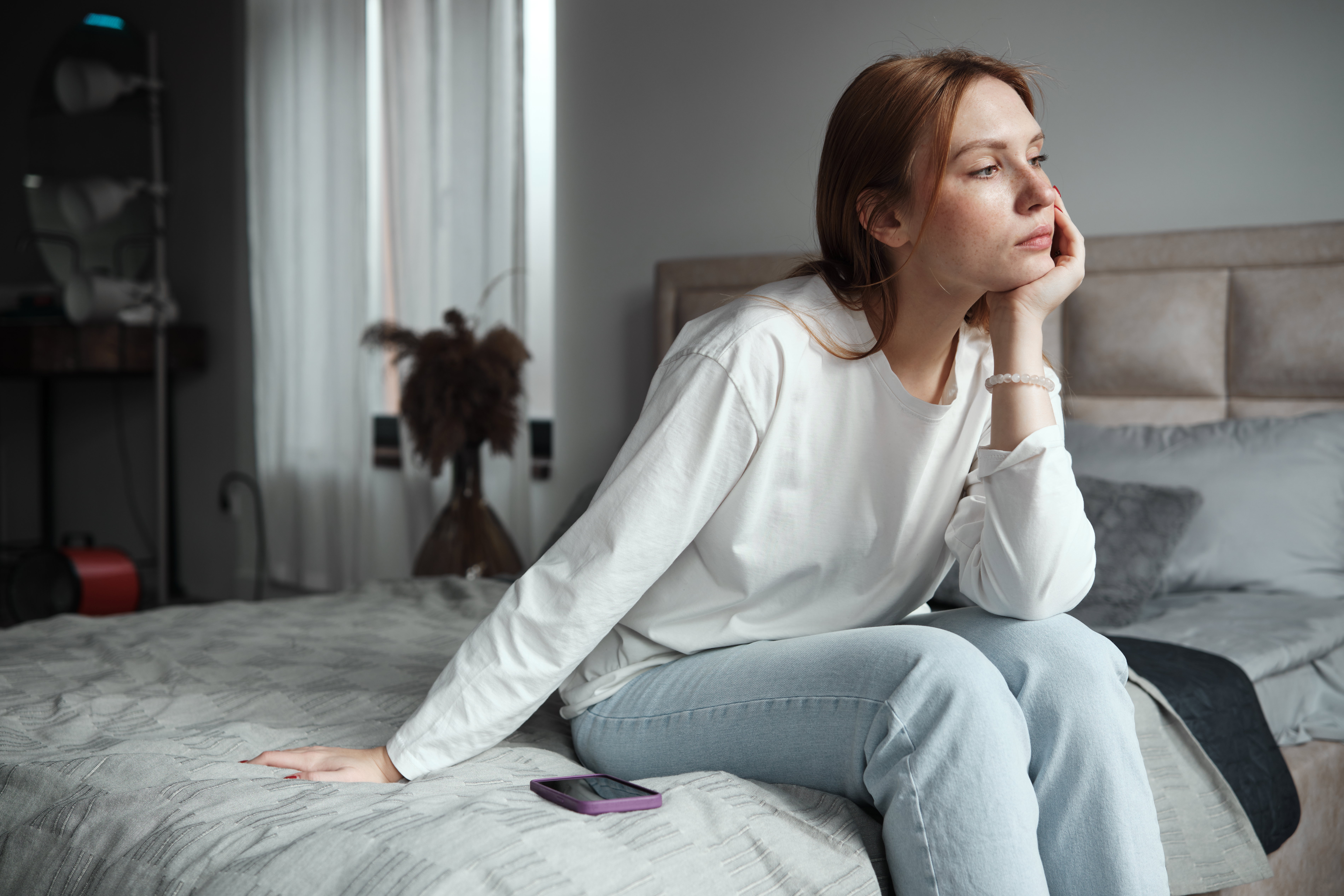 Phone anxiety. Upset woman sitting on bed with phone looking out window, feeling depressed, waiting for call. Social media depression