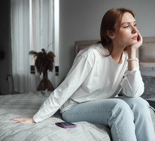 Phone anxiety. Upset woman sitting on bed with phone looking out window, feeling depressed, waiting for call. Social media depression