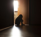 Despairing child sitting with head on knees in the dark frame of a doorway, backlit by a room behind flooded with daylight