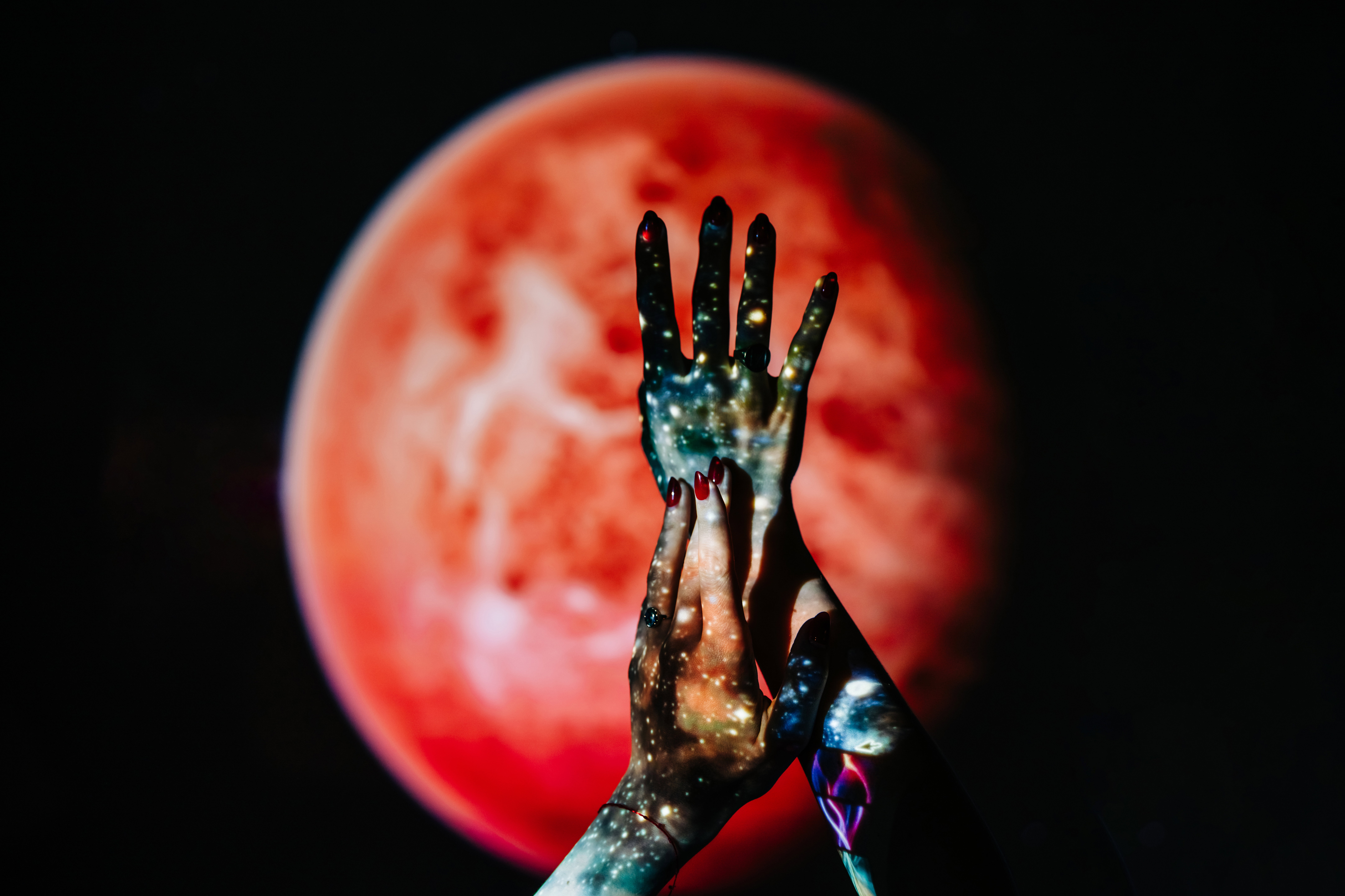 Hands with a galaxy projection are held up against a blurred red planet on a dark background. This artistic concept explores the connection between humanity, the cosmos, and inner space.