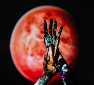 Hands with a galaxy projection are held up against a blurred red planet on a dark background. This artistic concept explores the connection between humanity, the cosmos, and inner space.