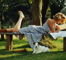 Woman lying on bench by tree reading, dog beneath bench