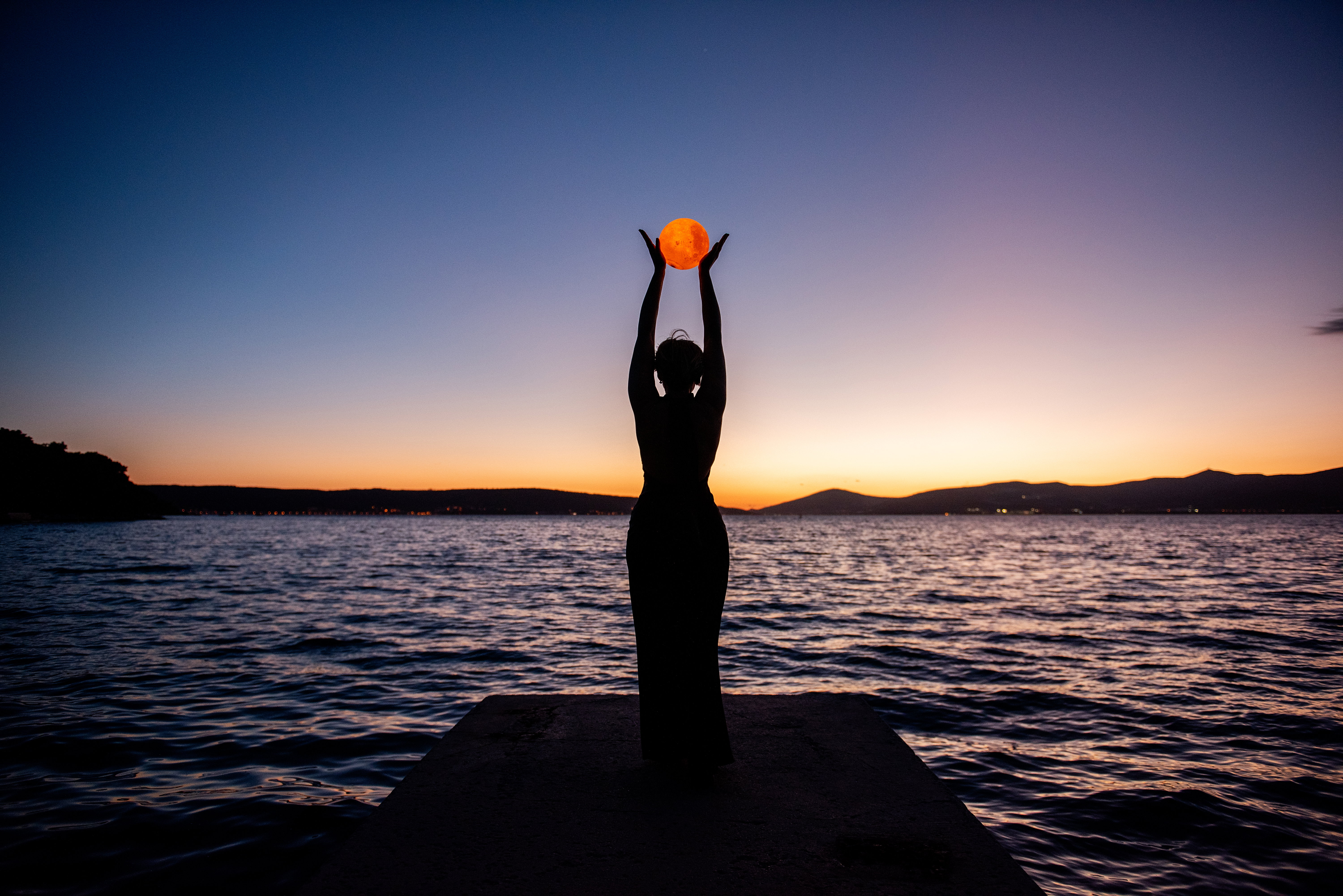 Silhouette of young woman in black dress, backdrop of twilight by sea, holding full moon in hands.