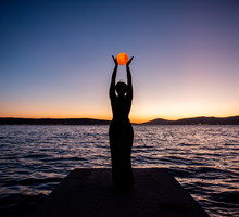 Silhouette of young woman in black dress, backdrop of twilight by sea, holding full moon in hands.