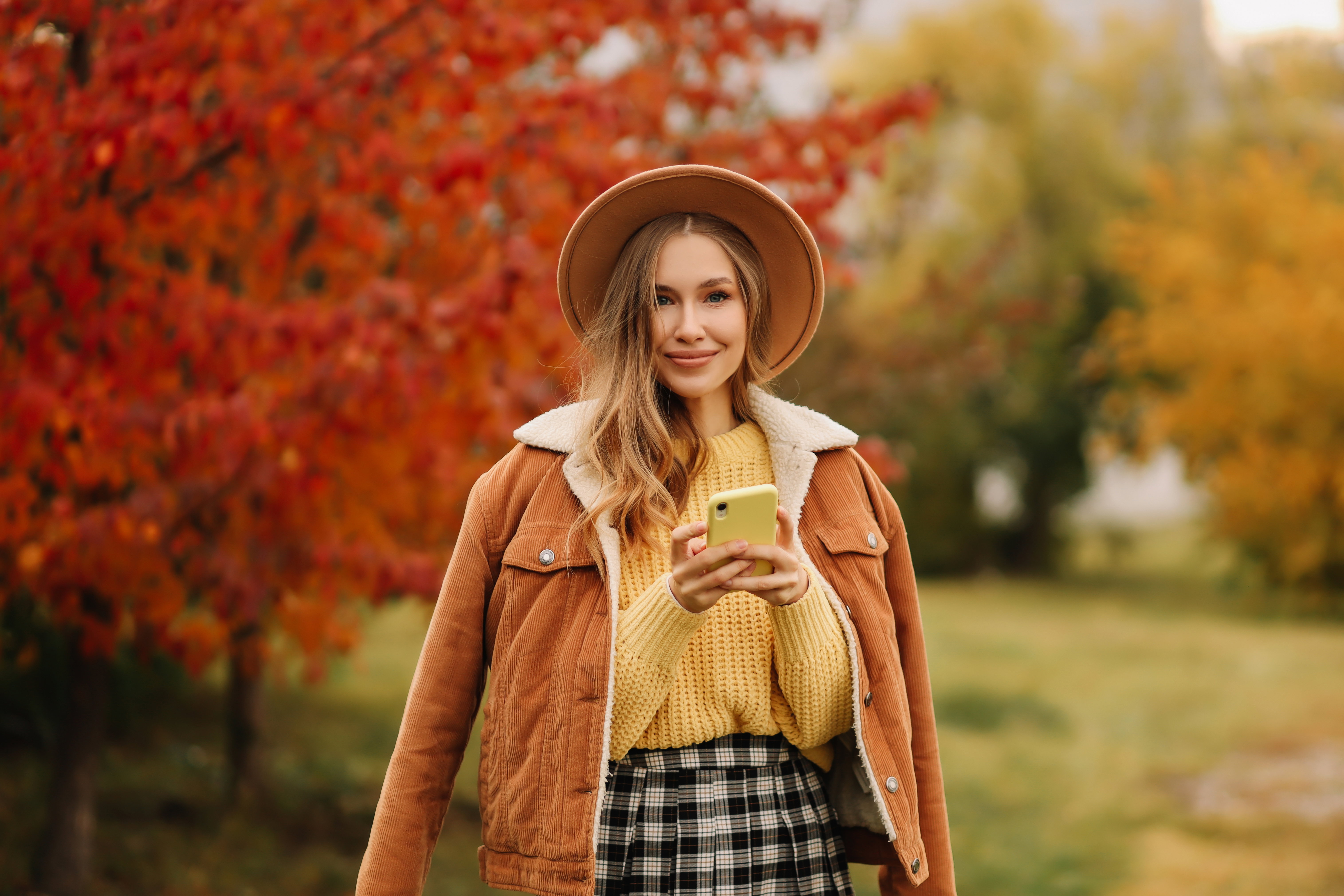 Portrait of a beautiful young woman of an influencer in warm clothes and a fashionable hat communicates online using a mobile phone and wireless technologies walking in an autumn park outdoors in autumn