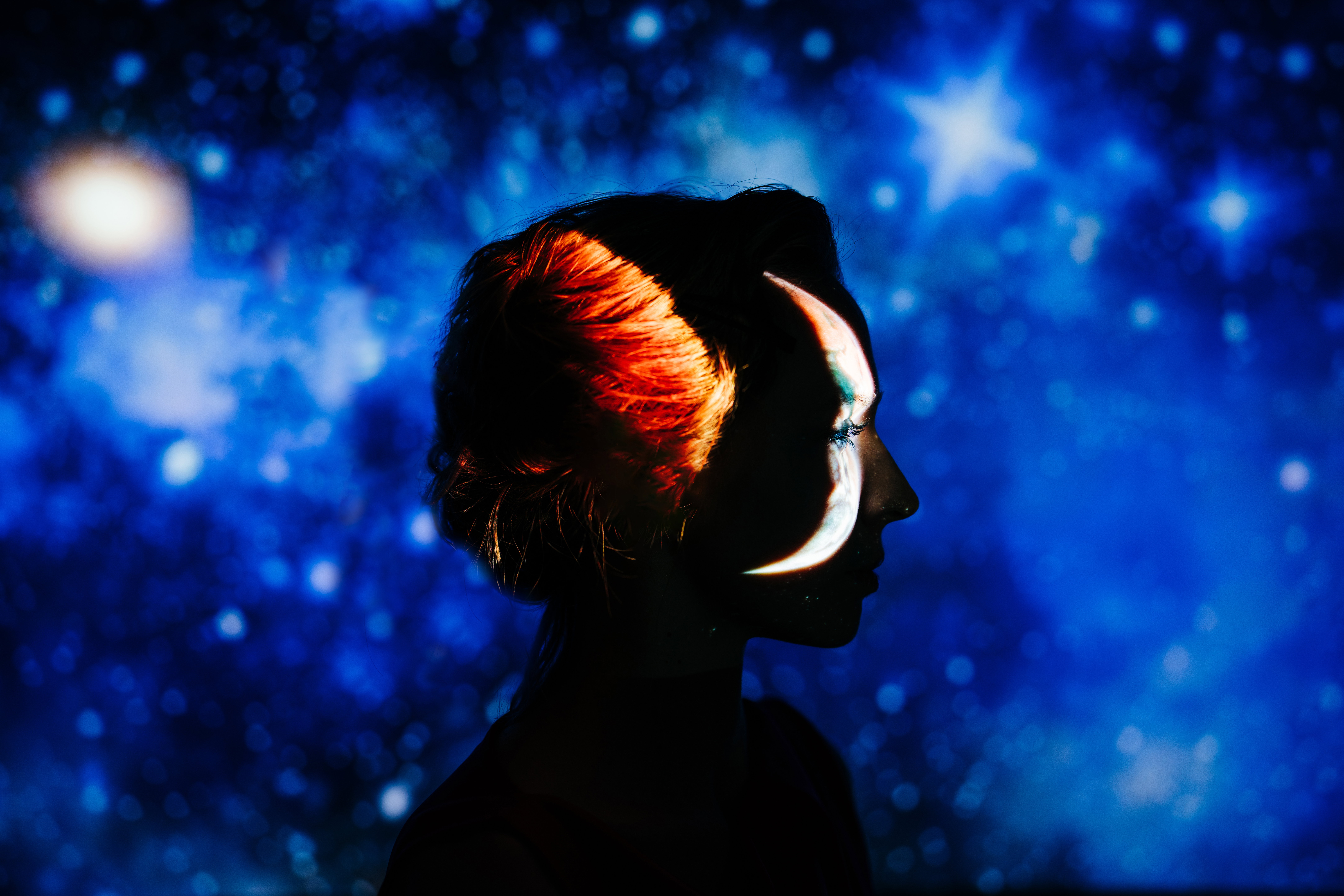 A striking profile portrait of a woman silhouetted against a deep blue starry night sky, with a crescent moon projected onto her face.