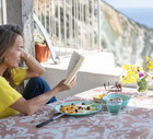 Woman enjoying a light lunch on a terrace with sea view, reading a book while eating