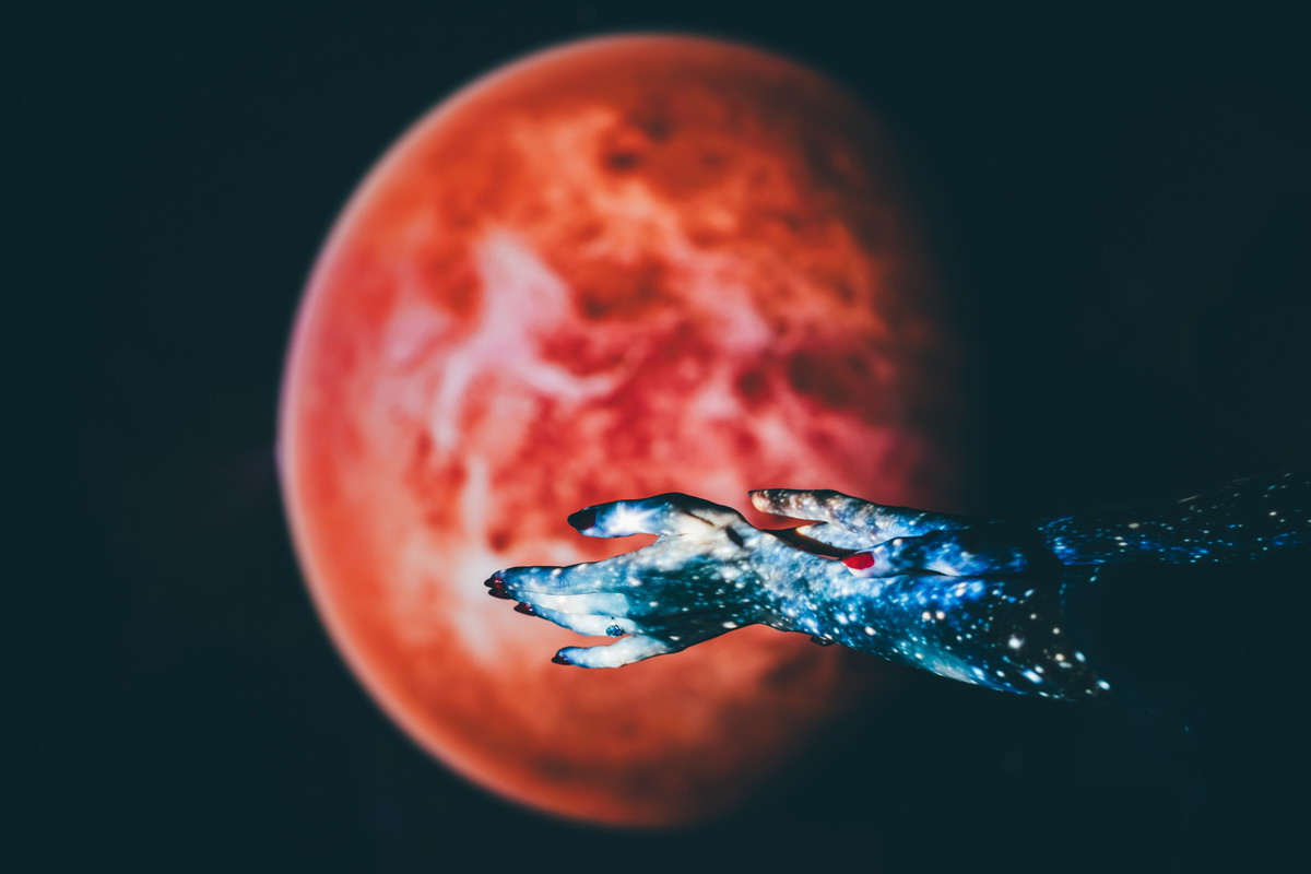 A close-up shot of two hands, projected with a vibrant blue galaxy pattern, are posed against a blurred red planet backdrop.