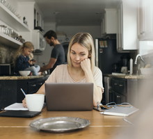 Mid adult woman using laptop at table with family in kitchen at home